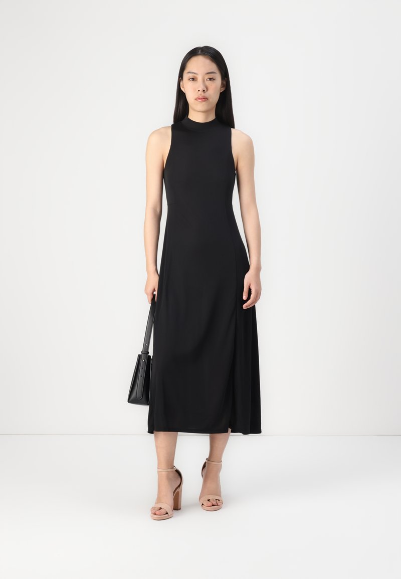 Schwarzes ärmelloses Midikleid mit hohem Ausschnitt, glatter Textur und seitlichem Schlitz. Kombiniert mit nuden Blockabsatzsandalen und einer schwarzen Handtasche.
