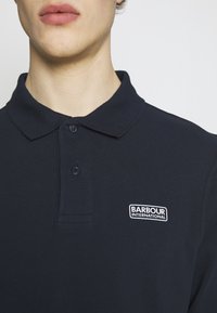 Młody mężczyzna w ciemnoniebieskiej koszulce polo z kołnierzykiem i logo "Barbour International" na lewej piersi.