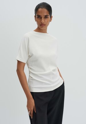 Frau mit zurückgebundenem dunklem Haar, die ein schlichtes weißes Kurzarmshirt und schwarze Hosen trägt, steht mit einer Hand in der Tasche.