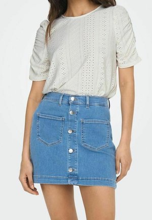 Vrouw draagt een wit ooglet blouse met korte mouwen, ingestopt in een blauwe denim minirok met knopen aan de voorkant en zakken.