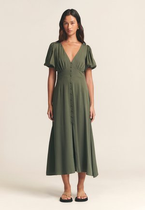 Olivově zelené midi šaty s nafouknutými rukávy, výstřihem do V, zapínáním na knoflíky a rozparkem ve spodní části; doplněné plochými sandály.