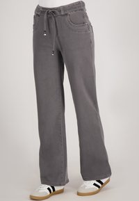 Grijze wijde broek met een trekkoord aan de voorkant en zakken, gedragen met witte sneakers met zwarte strepen en rubberen zolen.