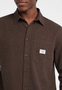 Camicia button-up a scacchi marroni con tasca, dotata di bottoni neri e un'etichetta di marca sul petto. Tessuto morbido e strutturato.