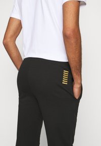 Svarta mjukisbyxor med en slät textur, som har en sidoficka och vertikala guldränder som designaccent. Midjebandet är elastiskt.