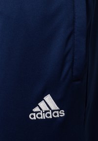 Tmavě modré sportovní kalhoty vyrobené z texturované látky, s bílou vyšívanou logem Adidas v levém dolním rohu. Viditelné dva boční kapsy.