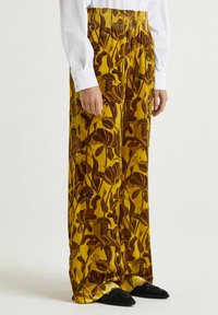 Pantaloni a gamba larga in tessuto giallo senape con motivo floreale marrone scuro. Caratterizzati da una cintura elastica e una texture morbida.