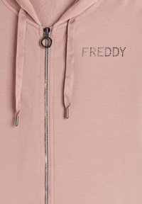 Felpa con zip di colore rosa chiaro, caratterizzata da una cerniera in tono argento, cordini testurizzati e dettagli in cristallo che formano la scritta "FREDDY" sul petto.