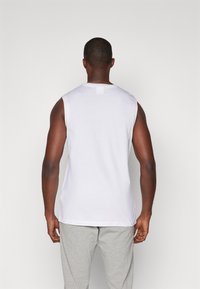 Witte mouwloze tanktop met een ronde halslijn, een gladde textuur en een rechte zoom, gedragen over grijze broek. Achteraanzicht laat schouder- en halsontwerp zien.