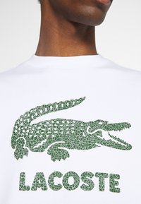 Sudadera blanca con un gran gráfico de cocodrilo verde con textura agrietada y la palabra "LACOSTE" en letras negritas debajo.