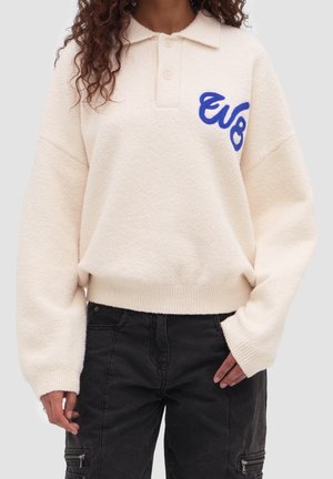 Personne portant un pull à manches longues couleur crème avec le logo bleu "evo" et un pantalon noir avec des poches zippées.