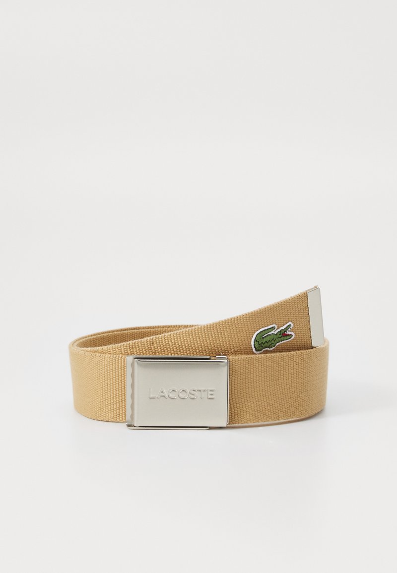 Cinturón de tela beige con una hebilla de metal plateada que presenta un logo grabado y un logo de cocodrilo bordado en verde en la correa.