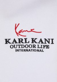 Tessuto bianco con testo ricamato. Firma rossa "Kane" sopra "KARL KANI" in grassetto nero, seguita da "OUTDOOR LIFE INTERNATIONAL" in nero standard.