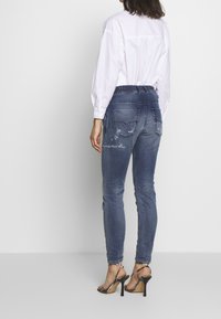 Nainen, joka on pukeutunut kuluneisiin sinisiin skinny-farkkuihin, valkoiseen pitkähiharaiseen paitaan ja mustiin korkokenkiin, seisoo yksinkertaista taustaa vasten.