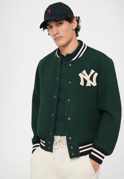 Tânăr îmbrăcat cu o jachetă varsity verde închis cu un logo alb NY, un polo verde închis, pantaloni de trening crem și o șapcă neagră cu emblemă roșie, cu mâinile în buzunare.