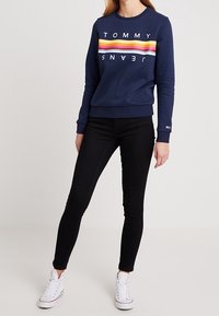 Kvinna iklädd en marinblå Tommy Jeans sweatshirt med flerfärgade ränder, svarta skinny jeans och vita högklackade sneakers som står mot en enfärgad bakgrund.