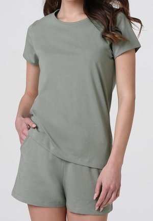 Donna che indossa un top a maniche corte verde salvia e pantaloncini abbinati, con una mano in tasca, in piedi davanti a uno sfondo semplice.