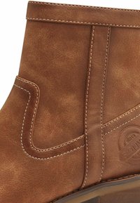 Bottine en cuir marron avec une texture lisse, des coutures contrastées, un détail de couture latérale et un accent logo circulaire sur le côté extérieur.