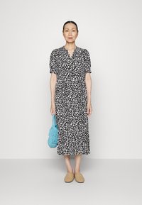 Vestido de estampa de leopardo em preto e branco, com mangas curtas e bufantes, decote em V e saia em camadas. Combinado com uma bolsa azul e chinelos bege.
