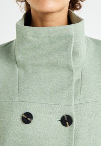 Abrigo color verde claro con cuello, que presenta una tela texturizada, doble botonera negra y un cuello estructurado. Vista en primer plano de los detalles.