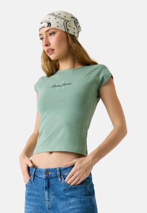 Giovane donna che indossa una t-shirt corta attillata verde chiaro e jeans blu con le mani nelle tasche, fazzoletto in testa con motivi di stelle e lune nere.