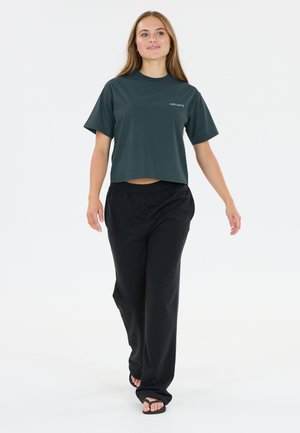 Ung kvinde gående iført en mørkegrøn cropped t-shirt med teksten "LES ARCS", sorte vidde bukser og sorte sandaler på hvid baggrund.