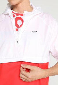 Personne portant une veste Nike F.C. blanche et rouge, en train de fermer la veste d’une main, vue partielle du cou à la taille.