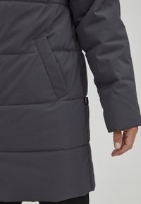 Mørkegrå polstret jakke med en glattere tekstur, har en sidelomme og et logomerke på faldet. Designet er langt og tætsiddende.