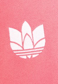 Růžová textilie s bílou vyšívanou logem se třemi okvětními lístky a vodorovnými pruhy pod ním, zdůrazňující hladkou pletenou strukturu.