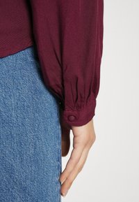 Main reposant à côté d'un jean en denim bleu, portant une chemise à manches longues bordeaux avec un poignet boutonné sur un fond blanc uni.