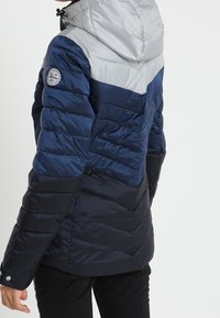 Person, der eine kapuzengefütterte, farblich abgestimmte Daunenjacke in Grautönen, Blau und Schwarz trägt, von hinten und von der Seite betrachtet.