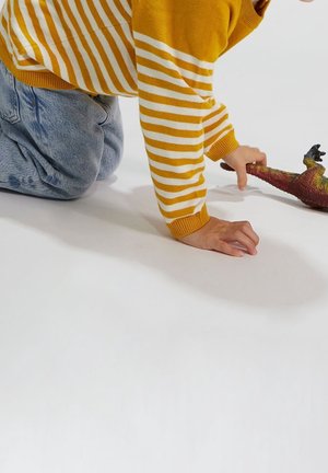Enfant portant un pull à rayures jaunes et blanches avec un jean, à genoux sur une surface blanche, tendant la main vers un jouet dinosaure aux couleurs vives.