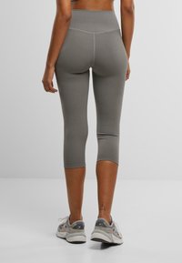 Grå leggings til midten av leggen med høy linning, glatt tekstur og sømdetaljer. Båret sammen med grå joggesko på en ensfarget bakgrunn.