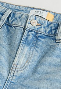 Vaaleansiniset bootcut-farkut, joissa on matala vyötärö, metallinen nappi, kontrastiväriset ompeleet ja haalistunut denim-tekstuuri.