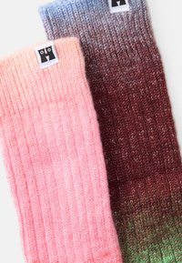Stickade benvärmare i mjuk rosa och gradientnyanser av röd till grön, med ribbad textur och liten rektangulär svart varumärkesetikett.
