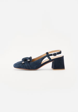 Marineblå ruskinds slingback-sko med kraftig mellemhøj hæl, side-rem med spænde, loafer-inspireret front og sølvfarvet dekorativt metal, på hvid baggrund.