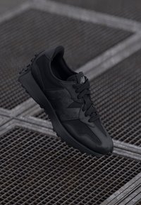 Crni New Balance tenisice s teksturiranom potplatom i mrežastim detaljima odmaraju se na metalnim rešetkama s isprepletenim linijama.