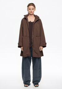 Chaqueta marrón de gran tamaño con capucha, cremallera delantera y dos bolsillos frontales, combinada con pantalones de chándal azules holgados y sandalias negras.