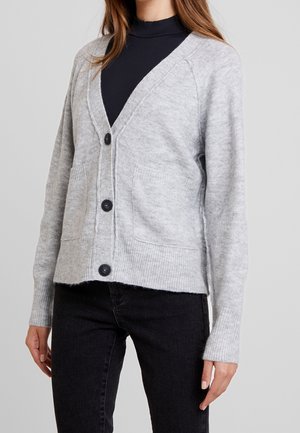 Femme portant un cardigan gris clair boutonné avec deux poches devant, par-dessus un haut noir côtelé et un pantalon noir.