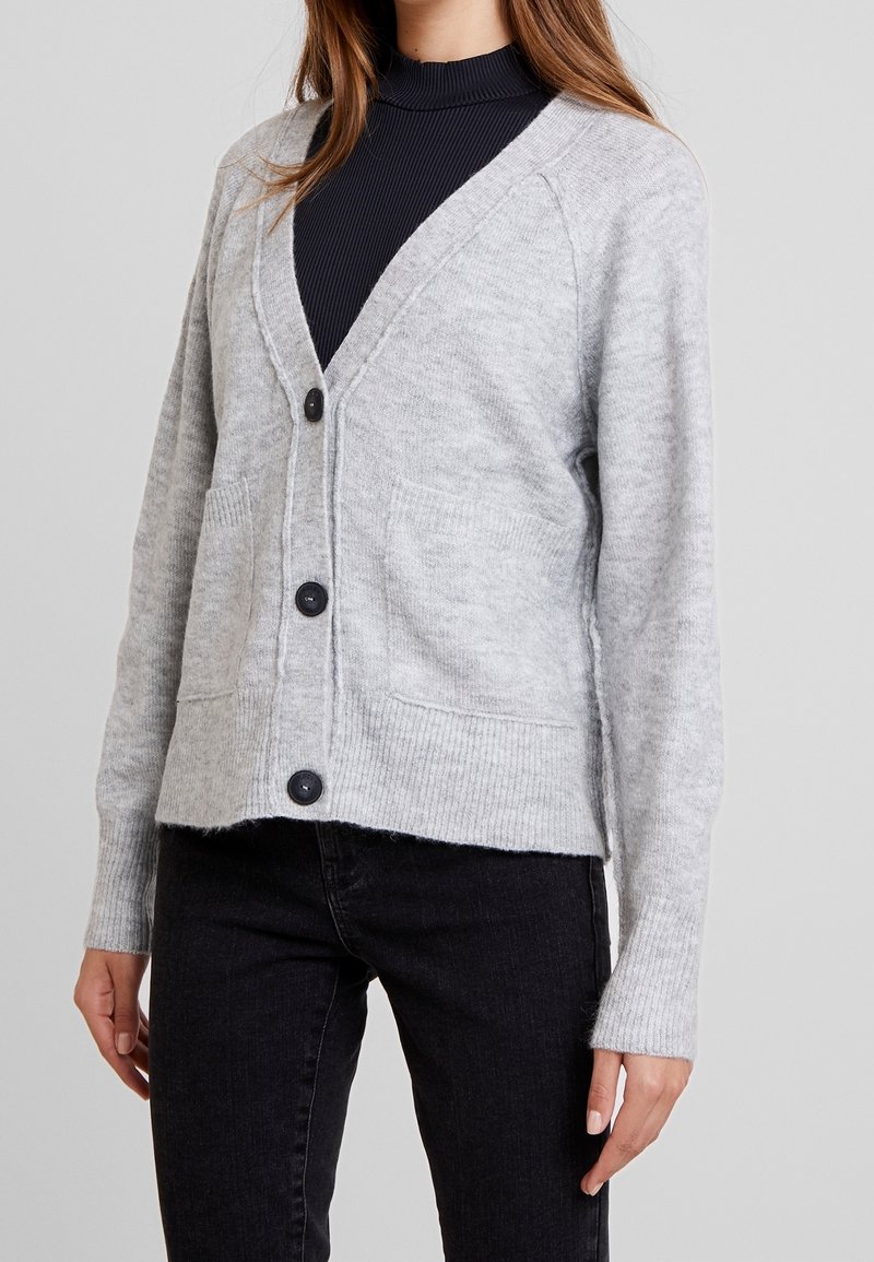 Femme portant un cardigan gris clair boutonné avec deux poches devant, par-dessus un haut noir côtelé et un pantalon noir.