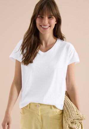 Donna sorridente con lunghi capelli castani che indossa una maglietta bianca e pantaloni gialli, che tiene una borsa di paglia intrecciata su uno sfondo beige.