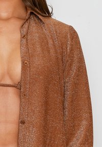 Camisa de bronce, con botones metálicos y un tejido texturizado y brillante. Presenta un cuello y un frente abierto, que revela un sujetador a juego.