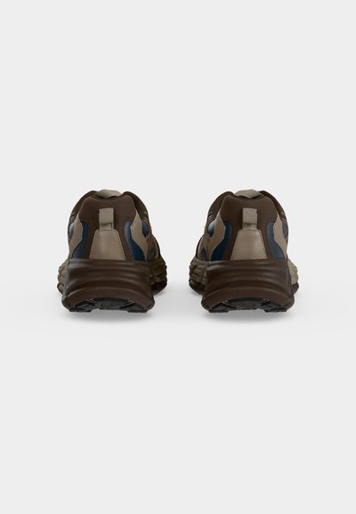 Paire de baskets marron et beige avec des semelles foncées vues de dos sur un fond gris clair uni.