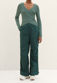 Zielony prążkowany top z kopertowym dekoltem w połączeniu z luźnymi, teksturowanymi ciemnozielonymi spodniami. Modelka ma na sobie białe sneakersy; oba elementy garderoby mają proste wzory.