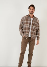 Camicia a quadri marrone e bianca con bottoni, indossata sopra una t-shirt bianca, abbinata a pantaloni marrone chiaro. Tessuto morbido e vestibilità casual.