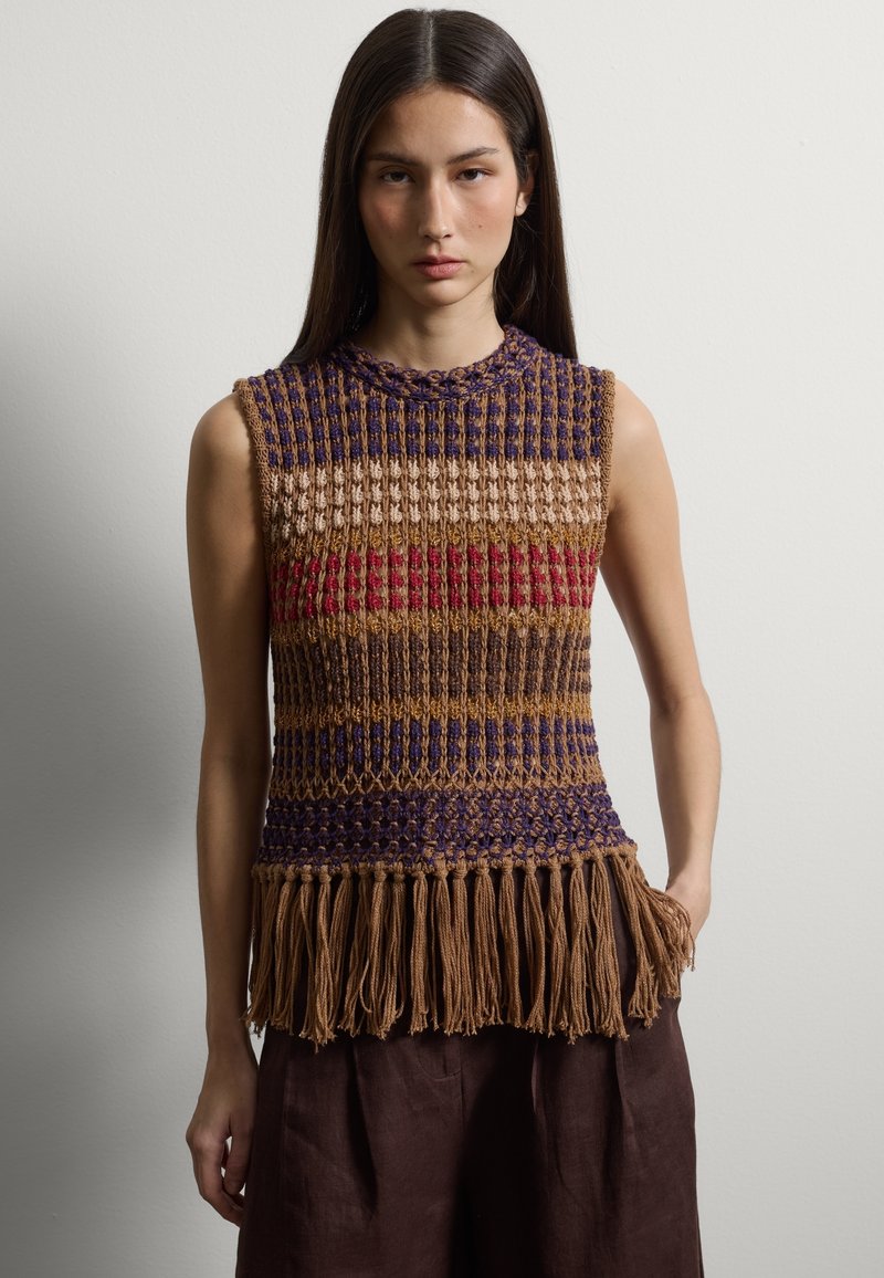 Junge Frau mit langem dunklem Haar, die ein ärmelloses, mehrfarbiges, gestricktes Oberteil mit Fransen am Saum und dunkelbraune Hosen trägt.