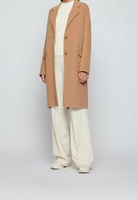 Cappotto lungo in misto lana cammello con rever, chiusura a singolo bottone, due tasche frontali e texture morbida, abbinato a un maglione e pantaloni color crema.