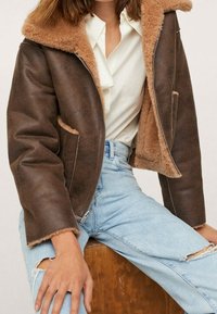 Femme portant une veste en peau retournée marron par-dessus une blouse blanche et un jean clair déchiré, assise avec les mains reposant sur les genoux.