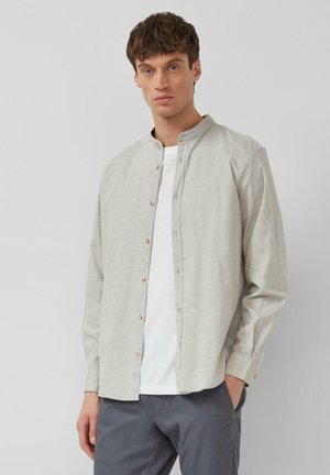 Junger Mann trägt ein hellgraues Hemd mit Stehkragen über einem weißen T-Shirt und dunkelgraue Hose, steht vor einfachem Hintergrund.