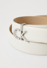 Witte leren riem met een gestructureerde oppervlakte en een zilverkleurige gesp met een gestileerd "CK"-logo. Glad interieur, ronde gespbevestiging.