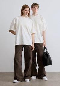 Λευκά υφασμάτινα μπλουζάκια συνδυασμένα με καφέ παντελόνια φαρδιάς γραμμής. Ένα άτομο κρατά μια μικρή μαύρη τσάντα. Και οι δύο φορούν λευκά αθλητικά παπούτσια και έχουν κοντά μαλλιά.